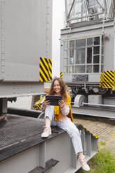 Glückliche junge Frau, die einen Tablet-PC auf dem Hafenkran eines Museums benutzt - IHF00962