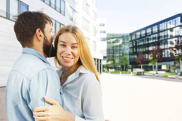 Cheerful blond woman with boyfriend standing in front of building - IHF00983