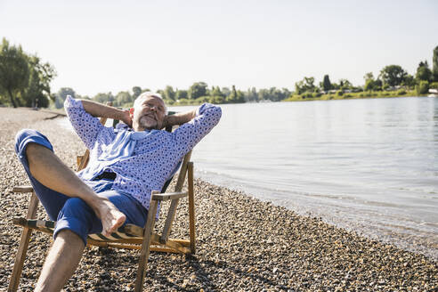 Smiling man with hands behind head resting on deck chair at riverbank - UUF26537