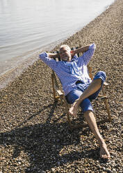 Smiling senior man with hands behind head resting on deck chair at riverbank - UUF26538