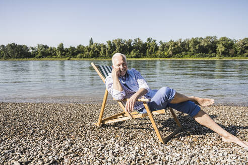 Happy retired man sitting on deck chair at riverbank - UUF26540