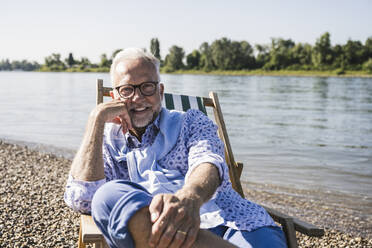 Smiling man wearing eyeglasses sitting on deck chair at riverbank - UUF26542