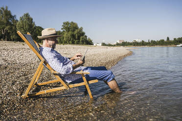 Smiling man using tablet PC sitting on deck chair at riverbank - UUF26549