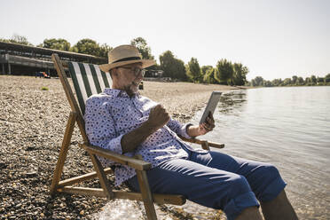 Happy man holding tablet PC sitting on deck chair at riverbank - UUF26550