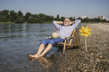 Lächelnder Mann mit den Händen hinter dem Kopf, der am Flussufer neben einem Papierspielzeug mit Windrädern sitzt - UUF26551