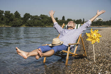 Verspielter älterer Mann mit erhobenen Armen, der am Flussufer neben einem Papierspielzeug mit Windrädern sitzt - UUF26552