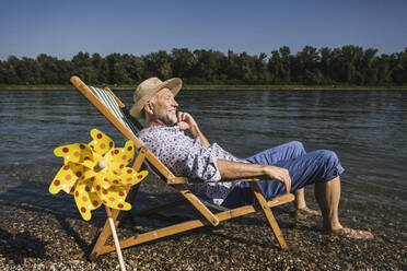 Lächelnder Mann, der am Flussufer neben einem Papierspielzeug mit Windrädern sitzt - UUF26563