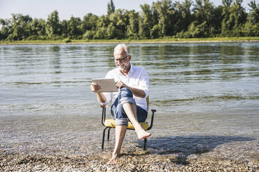Lächelnder Mann mit Tablet-PC auf einem Stuhl am Strand sitzend - UUF26566
