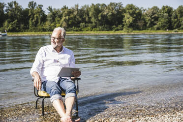 Älterer Mann mit Tablet-PC auf einem Stuhl am Flussufer sitzend - UUF26567