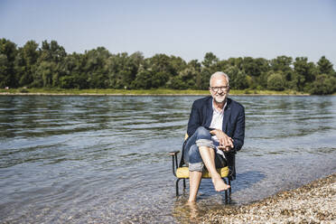 Lächelnder Mann mit Blazer sitzt auf einem Stuhl am Flussufer - UUF26568