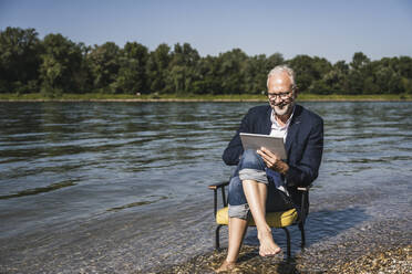 Lächelnder älterer Mann mit Tablet-PC auf einem Stuhl am Flussufer sitzend - UUF26569