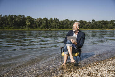 Man holding tablet PC sitting on chair at riverbank - UUF26570