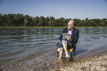 Mann mit Tablet-PC auf einem Stuhl am Flussufer sitzend - UUF26570