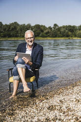 Lächelnder Mann mit Tablet-PC auf einem Stuhl am Flussufer sitzend - UUF26571