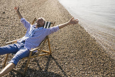 Happy senior man sitting with arms outstretched on deck chair at riverbank - UUF26584