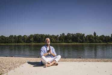 Senior man practicing yoga at riverbank on sunny day - UUF26606