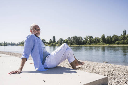 Älterer Mann mit gekreuzten Beinen am Flussufer an einem sonnigen Tag - UUF26608