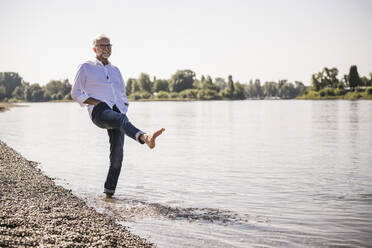 Lächelnder Mann mit Händen in den Taschen, der am Flussufer Wasser spritzt - UUF26618