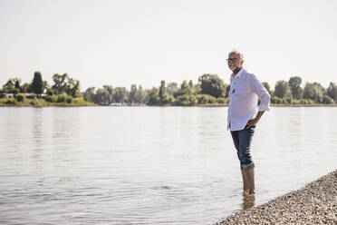 Lächelnder Mann, der mit der Hand auf der Hüfte am Flussufer steht - UUF26621