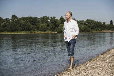 Älterer Mann mit Händen in den Taschen beim Spaziergang am Flussufer - UUF26623