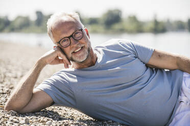 Smiling man lying at riverbank on sunny day - UUF26626