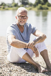Smiling man wearing eyeglasses sitting at riverbank - UUF26627