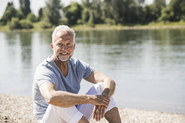 Lächelnder älterer Mann sitzt am Flussufer an einem sonnigen Tag - UUF26630