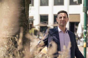 Smiling businessman standing near tree trunk - JOSEF10585