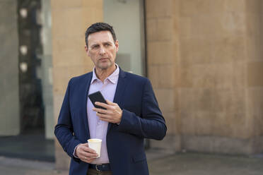 Contemplative businessman holding smart phone and disposable cup - JOSEF10589