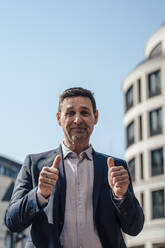 Smiling mature businessman showing thumbs up on sunny day - JOSEF10599