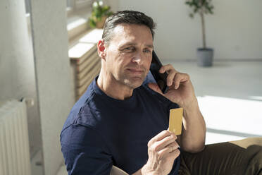 Lächelnder Geschäftsmann, der eine Kreditkarte hält und mit einem Smartphone im Büro telefoniert - JOSEF10610