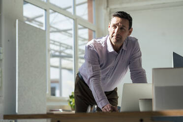 Nachdenklicher Geschäftsmann mit Laptop, der sich im Büro auf einen Tisch stützt - JOSEF10657