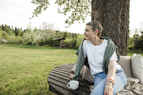 Smiling woman with coffee cup sitting on bench in front of tree trunk in garden - LLUF00697
