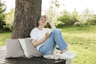 Smiling woman holding coffee cup sitting on bench around tree trunk - LLUF00701