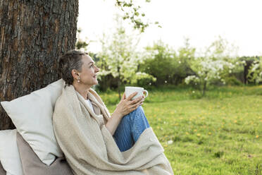 Happy woman wrapped in blanket holding cup sitting on bench around tree trunk - LLUF00706