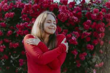 Smiling woman hugging self in front of rose bush - OSF00129