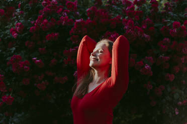 Glückliche Frau mit Händen hinter dem Kopf genießt das Sonnenlicht vor einem Rosenbusch - OSF00130