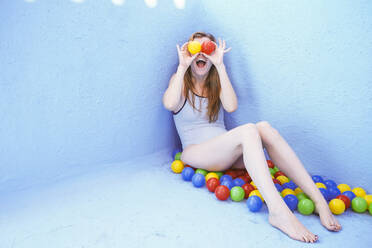 Happy woman covering eyes with balls sitting in front of blue pool wall - SVCF00102