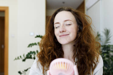Lächelnde Frau mit geschlossenen Augen, die einen tragbaren Ventilator zu Hause hält - TYF00302
