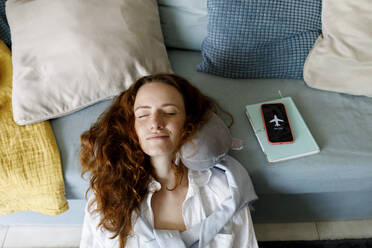 Smiling redhead woman leaning head by smart phone on sofa relaxing at home - TYF00325