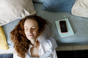 Lächelnde Frau mit Nackenkissen entspannt sich mit ihrem Smartphone auf dem Sofa zu Hause - TYF00326