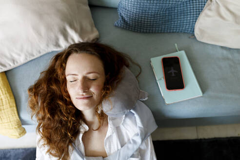 Smiling woman with neck pillow relaxing by smart phone on sofa at home - TYF00326