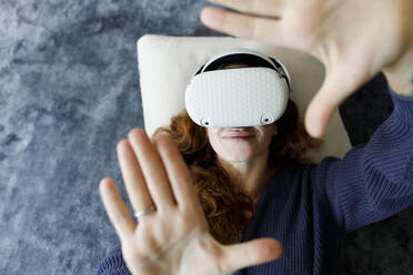 Glückliche Frau, die auf einem Teppich liegt und ein VR-Headset trägt, gestikuliert zu Hause - TYF00334