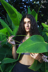 Teenager-Mädchen mit schwarzen Haaren steht bei einer Pflanze im Park - RFTF00263