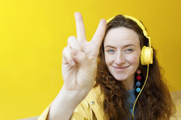 Lächelnde Frau mit Kopfhörern, die vor einer gelben Wand ein Friedenszeichen macht - TYF00346