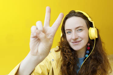 Lächelnde Frau gestikuliert mit Kopfhörern und hört Musik vor einer gelben Wand - TYF00347