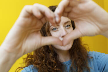 Woman gesturing heart shape in front of yellow wall - TYF00351