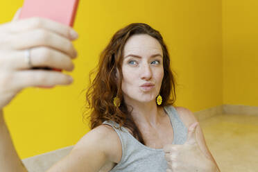 Frau gestikuliert Daumen hoch und macht ein Selfie mit ihrem Smartphone vor einer gelben Wand - TYF00353