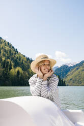 Smiling woman wearing hat enjoying boat ride in Lake Ritsa on sunny day - OMIF00894