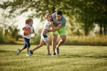 Fröhlicher Vater, der seiner Tochter hinterherläuft, die mit einem Rugbyball von einem Jungen auf einem Sportplatz gespielt wird - ZEDF04638